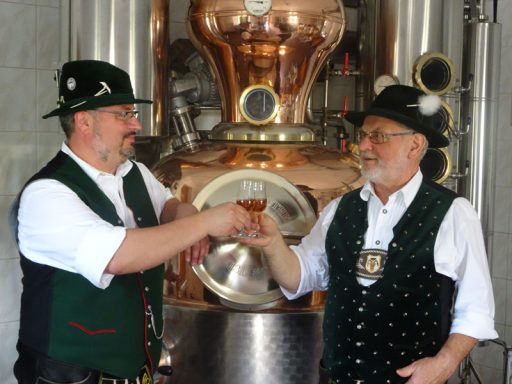 Rupert Vodermaier und Georg Vodermaier in Trachten stoßen mit einem Glas an, im Hintergrund eine Destille.