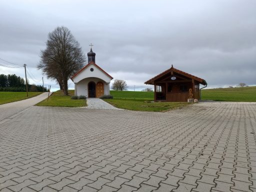 Kapelle und Holzunterstand auf einer Grünfläche mit gepflastertem Weg.