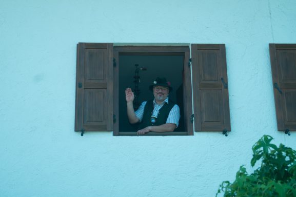 Rupert Vodermaier in traditioneller Kleidung winkt aus einem Fenster mit Holzladen.