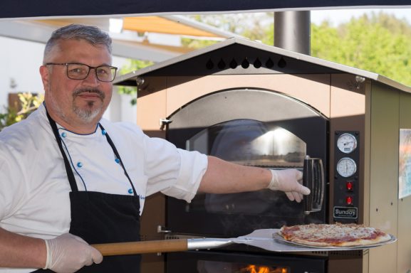 Rupert Vodermaier mit Brille hält eine Pizza vor einem Holzofen.