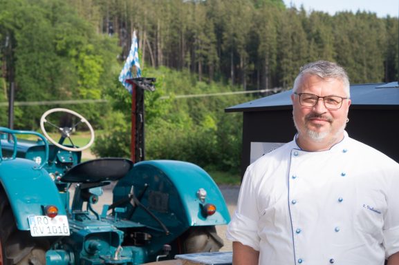 Rupert Vodermaier in Kochkleidung steht neben einem alten, türkisfarbenen Traktor im Freien.