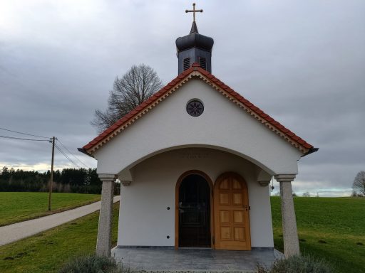 Kleine Kapelle mit Kreuz, umgeben von Wiesen und einer ruhigen Straße.